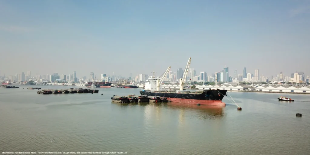Cargo ship anchored near a busy port with a modern city skyline in the background, illustrating maritime trade routes connected to the Hormuz Crisis discussed in a interview with Henrik Wachtmeister.